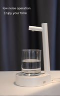 The mini water table dispenser in white on a desk in a bedroom, a glass cup half filled with water resting on the unit.