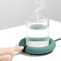 The green beverage warmer with a cup placed on top with steam evaporating, a hand and finger pressing the main button.